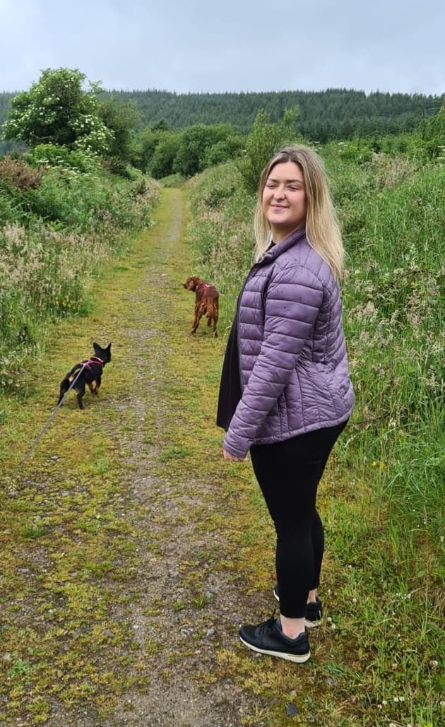 A woman in a purple jacket on a green grassy path with two dogs