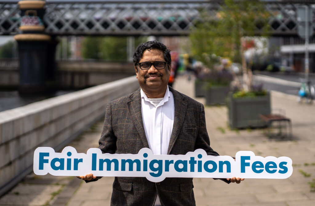 Senthil stands beside the River Liffey holding a sign that reads 'Fair Immigration Fees'