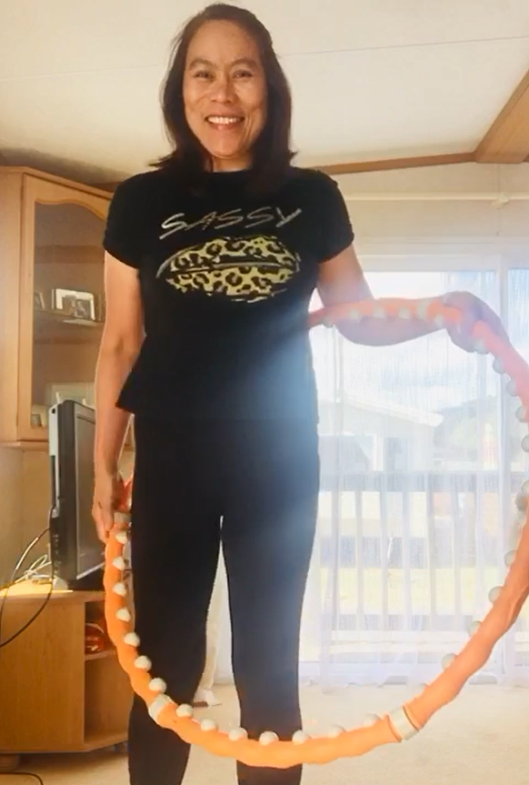 Mel stands in a sunlit room, smiling and holding a hula hoop.
